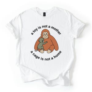 May include: White t-shirt featuring a cartoon orangutan and baby orangutan in shades of brown and green. The text "a toy is not a mother, a cage is not a home" is printed around the image in black.