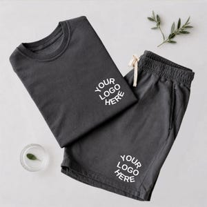 May include: A dark gray t-shirt and matching shorts set featuring the white text YOUR LOGO HERE printed on the chest of the shirt and the leg of the shorts. The shorts include a drawstring waistband and the set is paired with a small green leaf.