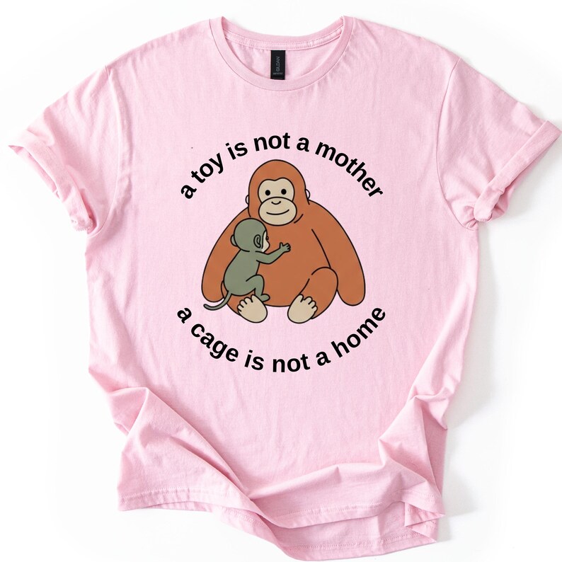 May include: A light pink t-shirt featuring a cartoon orangutan and baby ape illustration. The text "a toy is not a mother, a cage is not a home" is printed around the image in a circular pattern. The shirt has short sleeves.