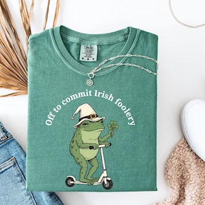 May include: A teal Comfort Colors t-shirt with a graphic of a frog wearing a leprechaun hat and riding a scooter, holding a four-leaf clover. The text reads "Off to commit Irish foolery." A silver necklace with a sun pendant is draped on the shirt.