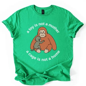May include: Green t-shirt featuring a cartoon orangutan embracing a baby orangutan. The text "a toy is not a mother, a cage is not a home" is arched around the image in white. The shirt has short sleeves and a crew neck.