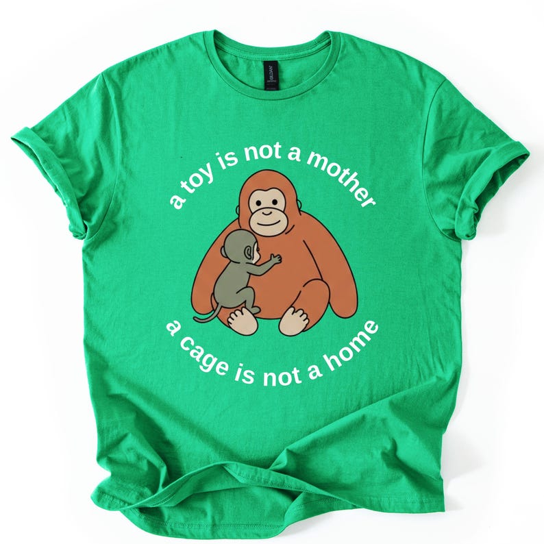 May include: Green t-shirt featuring a cartoon orangutan embracing a baby orangutan. The text "a toy is not a mother, a cage is not a home" is arched around the image in white. The shirt has short sleeves and a crew neck.