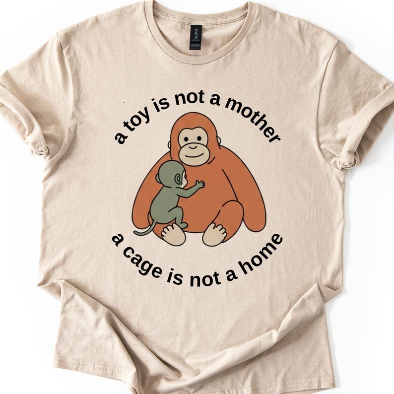 May include: Beige t-shirt featuring a cartoon orangutan embracing a smaller monkey. The text "a toy is not a mother, a cage is not a home" is printed around the image in black. The shirt has short sleeves and a crew neck.