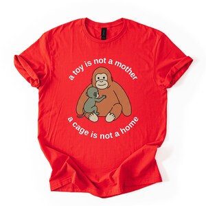 May include: Red t-shirt featuring a cartoon orangutan hugging a smaller ape, with the text "a toy is not a mother, a cage is not a home" in a circular design. The shirt is made of a soft material.
