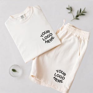 May include: A cream-colored t-shirt and matching shorts set featuring the black text "YOUR LOGO HERE" printed on the chest of the shirt and the lower leg of the shorts. The shirt has a crew neckline and the shorts feature an elasticized waistband.