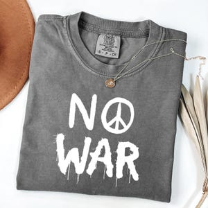 May include: A gray Comfort Colors t-shirt with the words "NO WAR" in white, featuring a peace symbol. A gold necklace with a pendant and a brown hat are visible. The shirt is folded on a white surface.