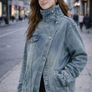 Könnte beinhalten: Eine hellblaue Jeansjacke mit hohem Stehkragen, asymmetrischer Knopfleiste mit silberfarbenen Metallknöpfen, langen Ärmeln und einer Brusttasche auf der rechten Seite. Die Jacke hat eine strukturierte Passform und deutlich sichtbare Nähte.