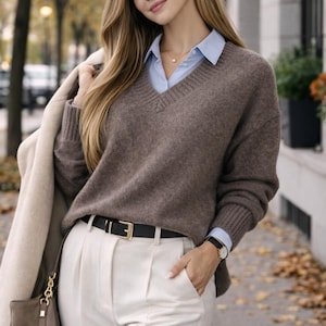Pode incluir: Uma mulher vestindo um suéter marrom com decote em V sobre uma camisa azul clara, combinada com calças brancas plissadas e um cinto preto com fivela dourada. Ela também está usando um relógio e segurando um casaco bege e uma bolsa marrom.