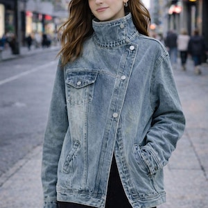 Può includere: Giacca di jeans blu chiaro con collo alto e chiusura asimmetrica con bottoni. La giacca ha due tasche frontali e viene indossata con pantaloni neri. La modella è in una via della città.