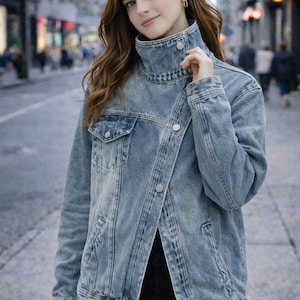 Könnte beinhalten: Hellblaue Jeansjacke mit hohem Kragen und Knopfverschluss. Die Jacke hat ein klassisches Design mit einer Vordertasche und einem leicht asymmetrischen Verschluss. Die Jacke wird über einem schwarzen Oberteil und einer schwarzen Hose getragen.