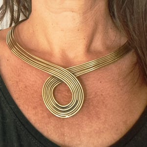 Op de afbeelding: Een goudkleurige metalen halsketting bestaande uit meerdere parallelle draden die in het midden samenkomen en een cirkelvormige lus vormen. De ketting rust rond de hals en heeft een gladde, gepolijste afwerking en een stijve vorm.