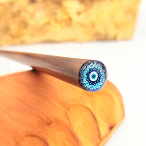 Peut inclure: Épingle à cheveux en bois marron avec une extrémité décorative ornée d'un motif d'œil bleu et blanc. Le motif de l'œil est entouré d'un motif floral. La tige est lisse.