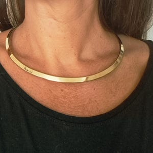 Puede incluir: Un collar tipo gargantilla de metal en tono dorado pulido con un diseño de banda plana, lisa y ligeramente curva. El collar se ajusta a la base del cuello y presenta un acabado reflectante de alto brillo que capta la luz de forma uniforme.
