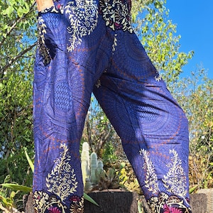 Può includere: Pantaloni blu dal taglio ampio con un motivo mandala ripetuto nei toni del blu scuro, crema e magenta. I pantaloni presentano una vita elastica arricciata, una silhouette rilassata e polsini elastici alle caviglie con tasche laterali.