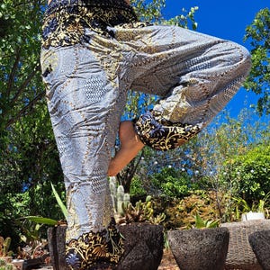 May include: Loose-fitting harem pants featuring a repeating mandala pattern in shades of grey, white, and black. The waistband and ankle cuffs are accented with a bold black and gold floral print, providing a distinct contrast to the main fabric.