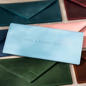 May include: A collection of colorful leather envelope-style wallets. The wallets are in various shades, including teal, navy, pink, and brown. One light blue wallet has the embossed text "FOR A RAINY DAY".