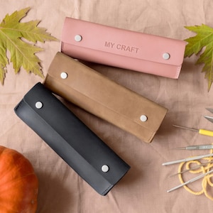 May include: Three leather craft tool cases in pink, brown, and black, each secured with two silver snaps. The pink case has "MY CRAFT" embossed on it. Crochet hooks, scissors, and a small pumpkin are also visible.