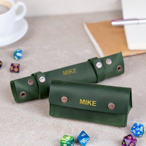 May include: Two green leather dice cases with the name "MIKE" in gold lettering. One case is rolled up, and the other is a small pouch. Several colorful polyhedral dice are scattered around the cases.