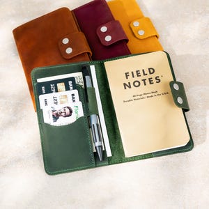 May include: Leather notebooks in shades of brown, burgundy, and yellow, with a green notebook open to reveal a "Field Notes" memo book, pen, and credit cards. The notebooks have snap closures.