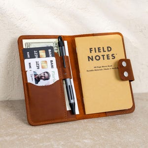 May include: Open brown leather wallet with a Field Notes memo book, credit cards, cash, and a pen. The memo book is tan with black text that reads "FIELD NOTES". The wallet has a snap closure.