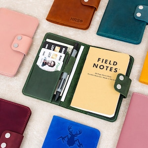 May include: A collection of leather notebooks in various colors, including green, brown, pink, and blue. One notebook is open, revealing a "Field Notes" memo book, a pen, and cards. The notebooks have button closures and embossed designs.
