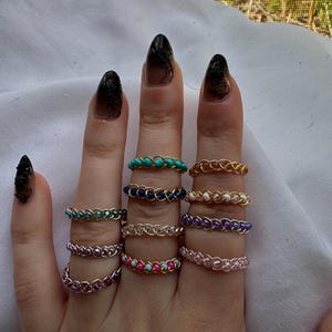 May include: A hand displaying a collection of wire-wrapped rings. The rings feature various colors of beads, including turquoise, blue, pink, and white. The wire is gold, silver, and copper-colored. The rings are worn on multiple fingers.