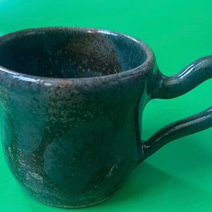 Può includere: Una tazza in ceramica con una smaltatura blu ottanio profondo e blu scuro con una texture maculata. La tazza presenta un corpo largo e arrotondato e un grande manico curvo che si estende dal lato, con una finitura lucida su tutta la superficie.
