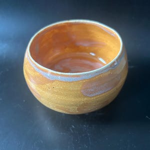 May include: A handmade ceramic bowl with a rounded shape. The bowl has a warm, earthy colour palette with shades of yellow, brown, and a hint of blue around the rim. The interior has a glossy finish.
