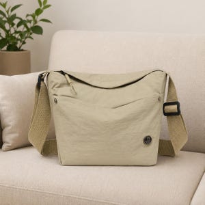 Op de afbeelding: Een beige messenger bag met een lange, verstelbare riem en een voorvak. De tas heeft een ritssluiting en een klein embleem op de voorkant. De tas is gemaakt van licht, gekreukt materiaal en ligt op een beige bank.