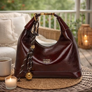 May include: A glossy, burgundy-colored handbag with a curved shape and a gold-toned handle. The bag features a decorative tassel and a gold-colored charm. The text "COIH ZDN" is visible on the front of the bag. The bag is sitting on a woven mat.