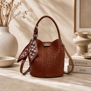 May include: A brown leather bucket bag with a woven texture, a top handle, and a detachable shoulder strap. A patterned scarf is tied to the handle. The bag has a small rectangular logo on the front.