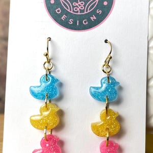 以下が含まれることがあります： 青、黄色、ピンクのグリッターレジンで作られたアヒルの形をした3つのチャームが付いた、ぶら下がりタイプのイヤリング。チャームはゴールドカラーのフープとフックで繋がれています。イヤリングは、丸いロゴが付いた白いカードに展示されています。