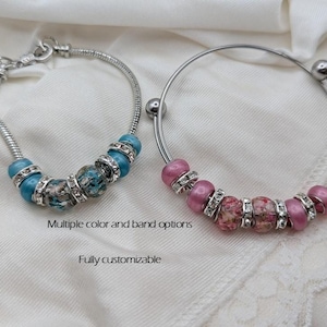 May include: Two bracelets with decorative beads. One bracelet is silver with blue and patterned beads, the other is a silver bangle with pink and floral beads. The text says "Multiple color and band options" and "Fully customizable".