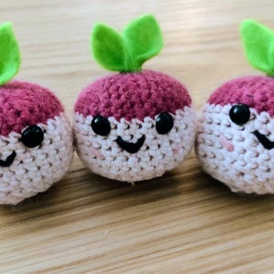 May include: Three crocheted radish toys with smiling faces. Each radish has a burgundy top, a white bottom, and green felt leaves. The radishes have black button eyes and a stitched black smile. They are arranged on a wooden surface.
