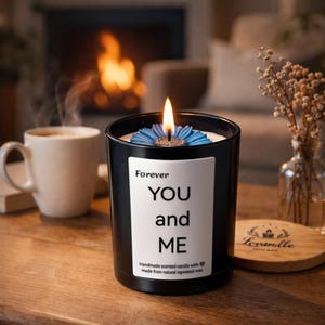 Puede incluir: Una vela negra encendida con un diseño de flor azul, con la etiqueta "Forever YOU and ME". La vela está sobre una superficie de madera junto a una taza de café y un pequeño jarrón con flores secas. La vela está hecha a mano con cera de colza natural.
