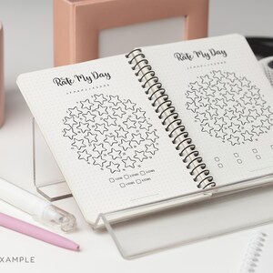 May include: Open spiral-bound notebook with the text "Rate My Day" and star-shaped rating charts. The notebook is open to two pages, each featuring a circular arrangement of stars for daily evaluation. Includes checkboxes for rating.