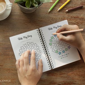May include: An open spiral-bound notebook with the text "Rate My Day" is shown. The notebook pages have star-shaped graphics for daily rating. A person is using a coloured pencil to fill in the stars. Coloured pencils and a cup of coffee are also visible.