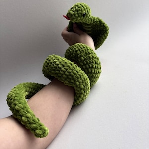 May include: A green crocheted snake toy with a red tongue. The snake is wrapped around an arm, with the head raised. The plush toy is made of a soft, textured yarn. The snake's body is a vibrant shade of green.