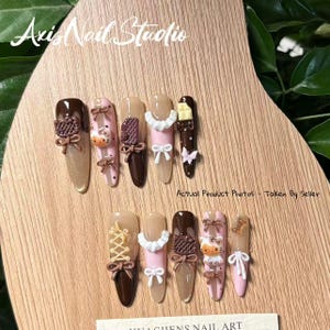 May include: A collection of almond-shaped acrylic nails with various designs. The nails feature brown, pink, and nude colors, adorned with bows, lace, and cartoon characters. The text "Axis Nail Studio" is visible at the top.