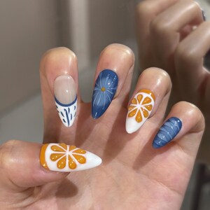 May include: Almond-shaped artificial nails featuring a variety of designs including white bases with raised orange citrus slice motifs, a solid blue nail with a white daisy illustration, a blue nail with white line patterns, and a French tip design.