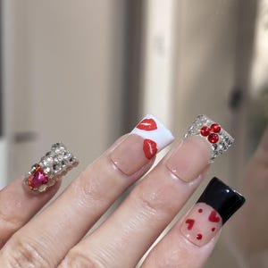 Pu&ograve; includere: Primo piano di una mano con unghie lunghe e decorate. Un'unghia presenta un gruppo di strass e una gemma rosa. Altre unghie hanno basi trasparenti con punte bianche, disegni di labbra e cuori rossi e punte nere con cuori rossi.