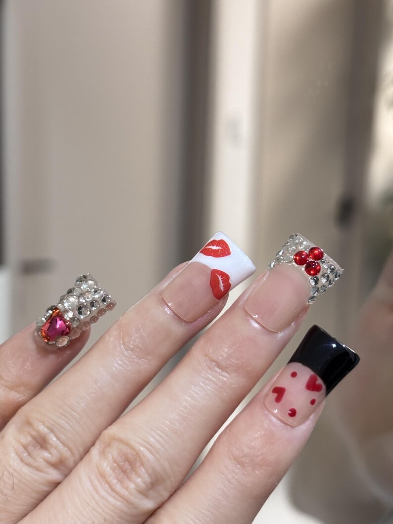 Pu&ograve; includere: Primo piano di una mano con unghie lunghe e decorate. Un'unghia presenta un gruppo di strass e una gemma rosa. Altre unghie hanno basi trasparenti con punte bianche, disegni di labbra e cuori rossi e punte nere con cuori rossi.