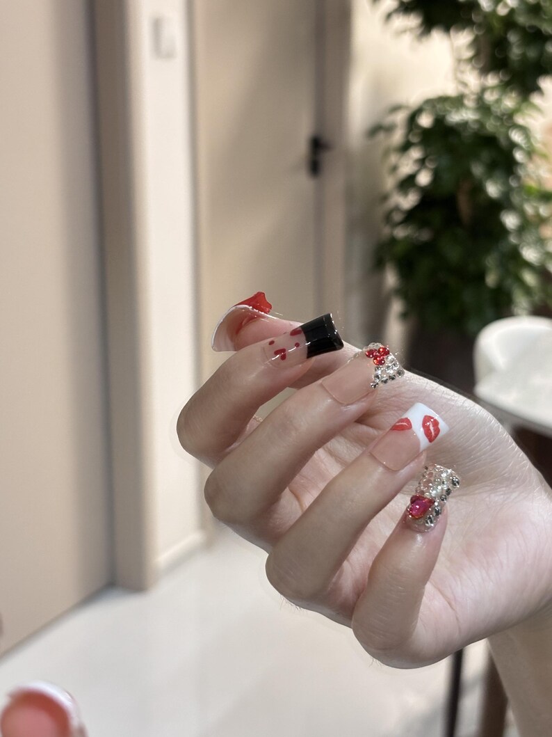 Pu&ograve; includere: Una mano con unghie lunghe e trasparenti in acrilico. Alcune unghie presentano disegni di rossetto rosso e nero, mentre altre sono decorate con strass scintillanti e gemme rosse. Le unghie hanno una forma quadrata e sono decorate con vari abbellimenti.