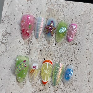 May include: A set of ten press-on nails featuring diverse designs including pink floral patterns, iridescent textures, a starfish, green tones with shell accents, citrus fruit slices, a jellyfish illustration, and blue floral embellishments.