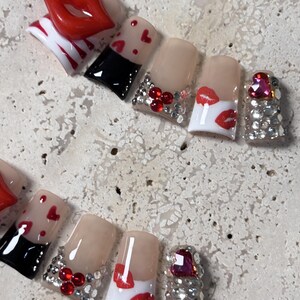 Pu&ograve; includere: Un set di unghie finte con vari disegni. Alcune unghie presentano labbra rosse, cuori rossi e strass. Un'unghia ha una grande gemma rosa a forma di cuore. Altre unghie hanno un design in bianco e nero.