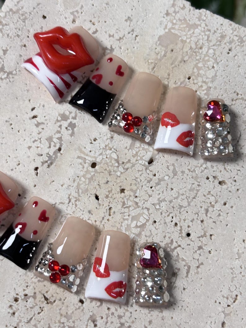 Pu&ograve; includere: Un set di unghie finte con vari disegni. Alcune unghie presentano labbra rosse, cuori rossi e strass. Un'unghia ha una grande gemma rosa a forma di cuore. Altre unghie hanno un design in bianco e nero.