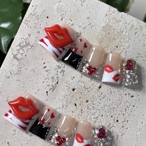 Pu&ograve; includere: Un set di unghie finte a tema San Valentino. Le unghie presentano labbra rosse, cuori e strass. Alcune unghie hanno un design in bianco e nero, mentre altre sono nude con accenti rossi.