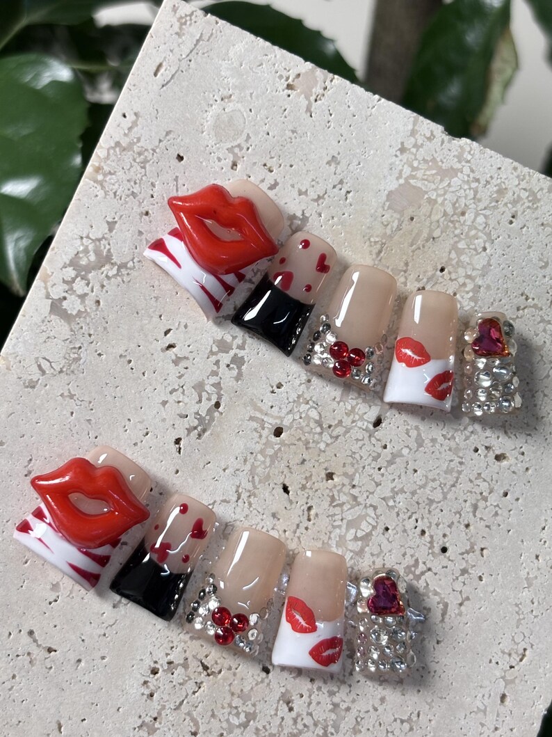 Pu&ograve; includere: Un set di unghie finte a tema San Valentino. Le unghie presentano labbra rosse, cuori e strass. Alcune unghie hanno un design in bianco e nero, mentre altre sono nude con accenti rossi.