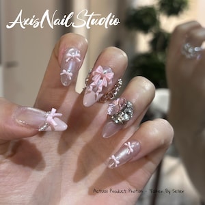 Puede incluir: Primer plano de una mano con uñas acrílicas largas en forma de almendra. Las uñas están pintadas en un diseño rosa claro y transparente, adornadas con pequeños lazos rosas y detalles plateados. Una uña presenta una gran gema rosa y un anillo plateado.