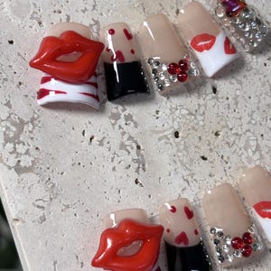 Pu&ograve; includere: Un set di unghie finte con vari design. Alcune unghie presentano labbra rosse, cuori rossi e una base nera. Altre sono decorate con strass e un top coat trasparente. Le unghie sono esposte su una superficie chiara e strutturata.